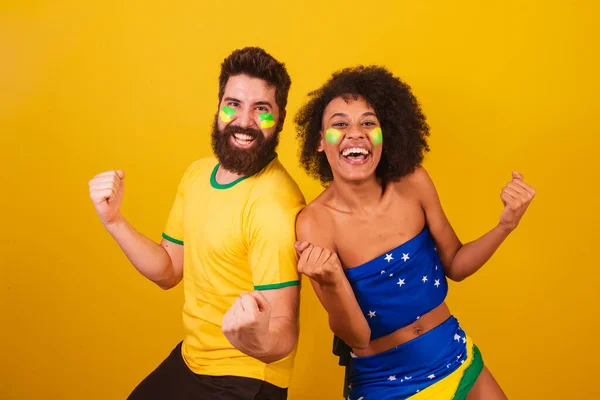 stock image couple of brazilian soccer fans, dressed in the colors of brazil, black woman, caucasian man. screaming goal