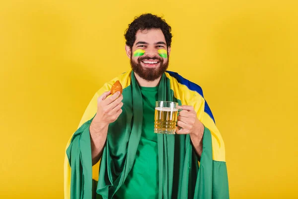 ブラジル出身の髭とブラジル人サッカーファンを持つ白人男性ビールとチキンドラムスティックのグラスを持ち典型的なブラジル料理 — ストック写真