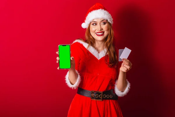 美丽的巴西红头发女人 穿着圣诞服装 圣诞老人 持有带有绿色彩色屏幕的信用卡和智能手机 — 图库照片