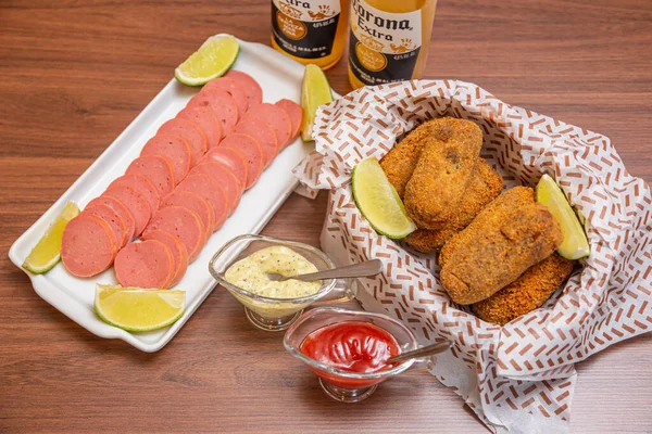 Chicken croquette and Cambu sausage,typical Brazilian snack, served with lemon slices, chili sauce and mayonnaise. On wooden table served with famous corona beer.