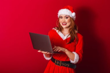 Güzel Brezilyalı kızıl saçlı kadın, Noel kıyafetleri giymiş, Noel Baba. dizüstü bilgisayarı tutuyor.