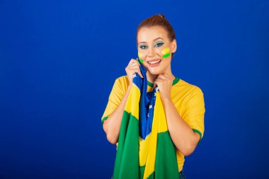 Beyaz kadın, kızıl saçlı, Brezilyalı futbol fanatiği, mavi arkaplan, elinde bayrakla gülümsüyor ve mutlu.