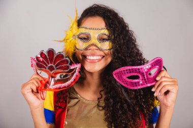 Güzel siyah Brezilyalı kadın, açık renkli elbiseli, karnaval. Maske takmak, karnaval maskeleri tutmak, insanları partiye davet etmek..