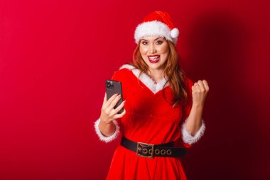Güzel Brezilyalı kızıl saçlı kadın, Noel kıyafetleri giymiş, Noel Baba. Akıllı telefonla kutlama.