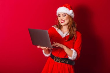 Güzel Brezilyalı kızıl saçlı kadın, Noel kıyafetleri giymiş, Noel Baba. dizüstü bilgisayarı tutuyor.