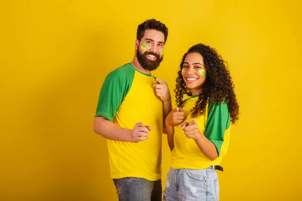 couple of brazil soccer supporters, dressed in the colors of the nation, black woman, caucasian man. Pointing at camera.