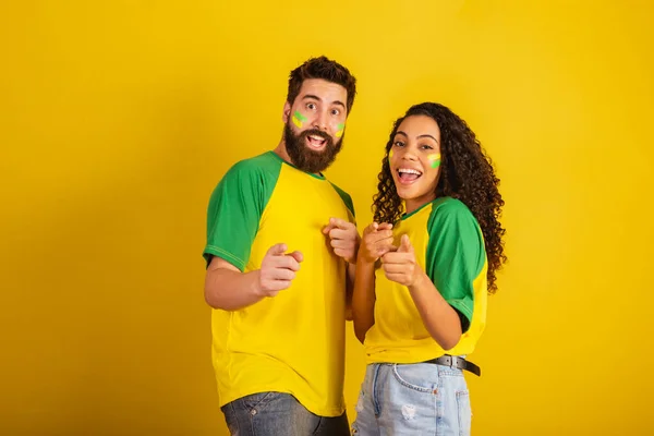 stock image couple of brazil soccer supporters, dressed in the colors of the nation, black woman, caucasian man. Pointing at camera.