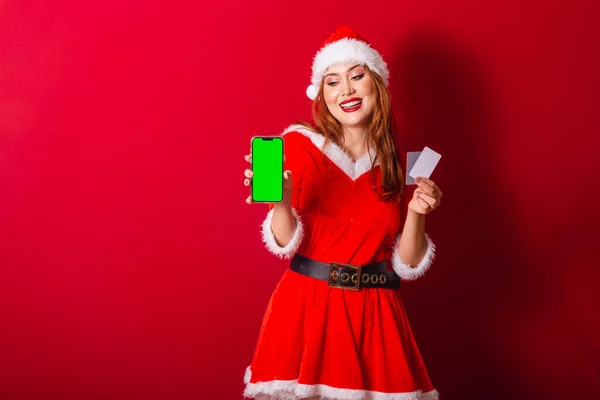 美丽的巴西红头发女人 穿着圣诞服装 圣诞老人 持有带有绿色彩色屏幕的信用卡和智能手机 — 图库照片