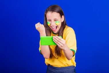 Genç bir kız, Brezilyalı bir futbol taraftarı. Cep telefonu tutmak, maç izlemek, Smartphone için tezahürat yapmak. uygulamalar.