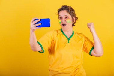 Yetişkin bir kadın, Brezilya futbolu fanatiği, akıllı telefon, cep telefonuyla futbol maçı izleyen,.