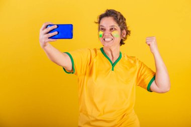 Yetişkin bir kadın, Brezilya futbolu fanatiği, akıllı telefon, cep telefonuyla futbol maçı izleyen,.