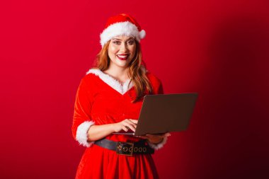 Güzel Brezilyalı kızıl saçlı kadın, Noel kıyafetleri giymiş, Noel Baba. dizüstü bilgisayarı tutuyor.