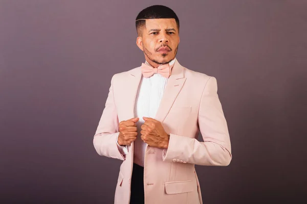 Brazilian black man, with pink suit and bow tie, gray background, holding suit