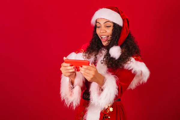 美丽的巴西黑人女人 穿着圣塔克洛斯的衣服 圣诞服装 用智能手机庆祝某事 — 图库照片