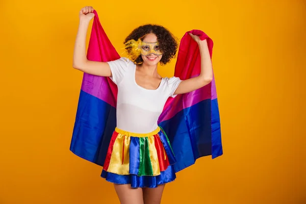 stock image Beautiful Brazilian woman, dressed in carnival clothes, colorful skirt and white shirt. wearing mask, holding bisexual flag.