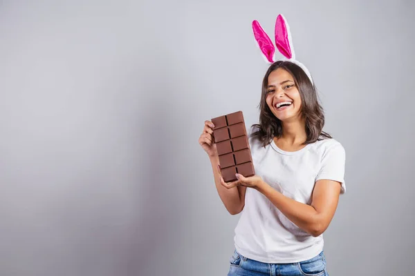 巴西女人 带着复活节兔子的耳朵 快乐地微笑着 手里拿着复活节巧克力棒 — 图库照片