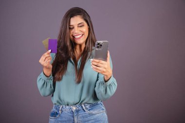 Yatay fotoğraf. Güzel Brezilyalı kadın, günlük kıyafetler, kot pantolon ve yeşil gömlekli. akıllı telefon ve kartlar, uygulama alımları, cep telefonları.