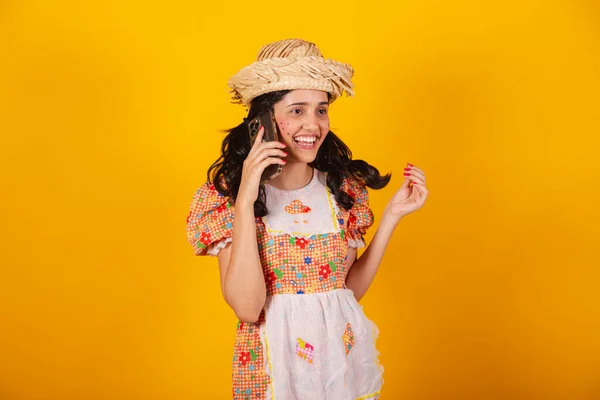 Beautiful Brazilian woman, with June party clothes, talking on voice call by smartphone.