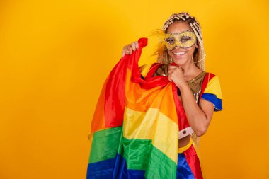 Güzel siyah Brezilyalı kadın, karnaval kıyafetleri giymiş, maske takıyor. Sarı arka plan. lgbt bayrağı tutuluyor.