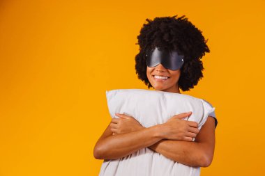 Uyuyor. Rüyalar. Kadın portresi. Pijamalı ve uyku maskeli Afro-Amerikalı bir kız, sarı bir arka planda yastığa sarılıyor ve gülümsüyor.