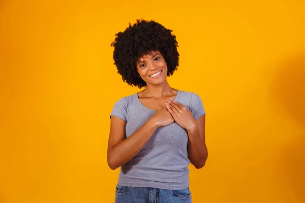 Grateful hopeful happy black woman holding hands on chest feeling pleased thankful, sincere african lady expressing heartfelt love appreciation gratitude honesty isolated on yellow studio background