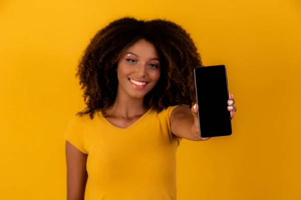 afro woman with curly hair pointing at cellphone on yellow background