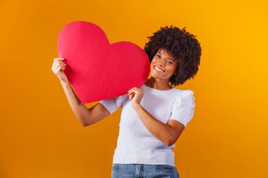 Elinde kocaman kırmızı bir kalp kartıyla gülümseyen afro bir kadının fotoğrafı.