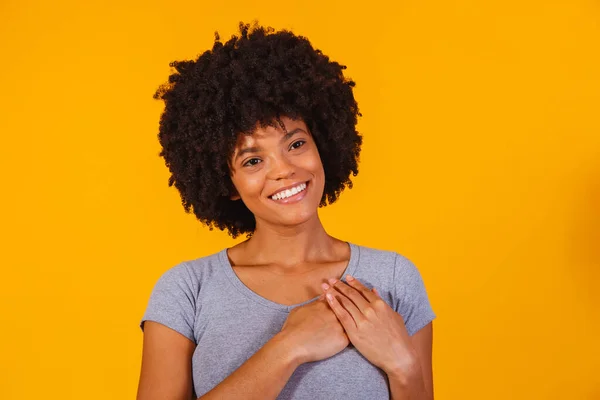Grateful hopeful happy black woman holding hands on chest feeling pleased thankful, sincere african lady expressing heartfelt love appreciation gratitude honesty isolated on yellow studio background