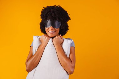 Uyuyor. Rüyalar. Kadın portresi. Pijamalı ve uyku maskeli Afro-Amerikalı bir kız, sarı bir arka planda yastığa sarılıyor ve gülümsüyor.