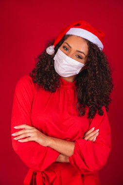 Noel Baba şapkalı genç bir kadın Covid-19 Coronavirus virüsü takıyor. Kırmızı arka plan stüdyosunda izole edilmiş maskeyi kurtardı. Mutlu Yeni Yıl Kutlaması Tatil Konsepti.