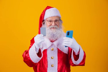 Yakışıklı, şaşkın sakallı Noel Baba 'nın portresi elinde plastik kart aygıtı web uygulama mağazası sipariş teslimatı ödemesi sarı arka planda izole edilmiş kablosuz ödeme.