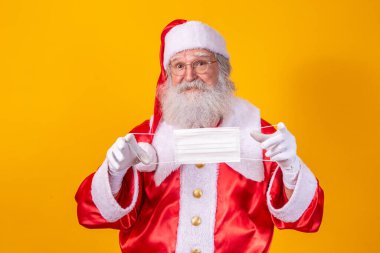 Sarı arka planı olan, koruyucu maskeli, gözlüklü ve şapkalı gerçek Noel Baba. Sosyal mesafeli bir Noel. Covid 19