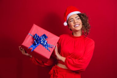 Noel Baba şapkalı genç afro kadın portresi kırmızı arka planda bir hediye taşıyor. Noel gecesi konsepti