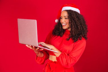 Kırmızı arka planda dizüstü bilgisayar kullanan Noel Baba şapkalı afro bir kadın..