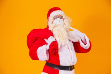 Sarı arka planda gerçek Noel Baba, Covid19 'a karşı koruyucu maske takıyor. Sosyal mesafeli bir Noel. Covid 19