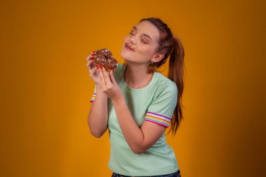 Güzel kızıl saçlı kız sarı arka planda çikolatalı donut yiyor. Kız donut yiyor.