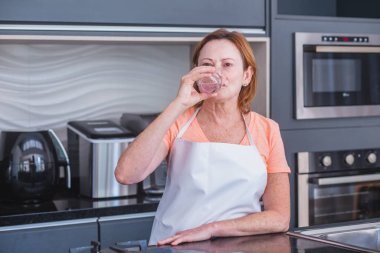 Olgun bir kadın sabah mutfakta su içiyor.
