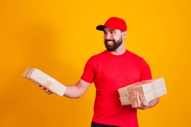 En iyi servis konsepti. Kırmızı şapkalı gülümseyen beyaz teslimatçı ve sarı stüdyo arka planında izole bir şekilde duran karton kutuları istifliyor. Posta taşıma paketleri