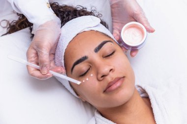 Yüzünde krema ve yanında kavanoz olan güzel bir kadının yakın çekimi. Yüz kremi süren bir kadın. spa konsepti