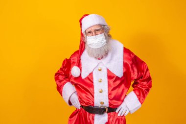 Sarı arka planda, koruyucu maske, gözlük ve şapka takan gerçek bir Noel Baba. Sosyal mesafeli bir Noel. Covid 19