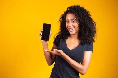 Afro saçlı, boş cep telefonu tutan ve sarı arka planı işaret eden mutlu, güzel bir kadın..