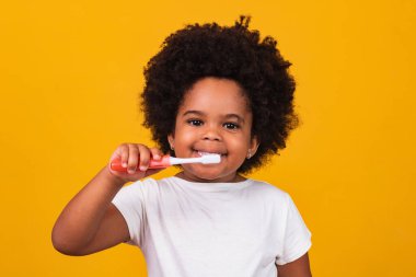 Dişlerini fırçalayan küçük tatlı bir Afrikalı Amerikalı kız, sarı arka planda izole edilmiş. Sağlıklı diş konsepti.