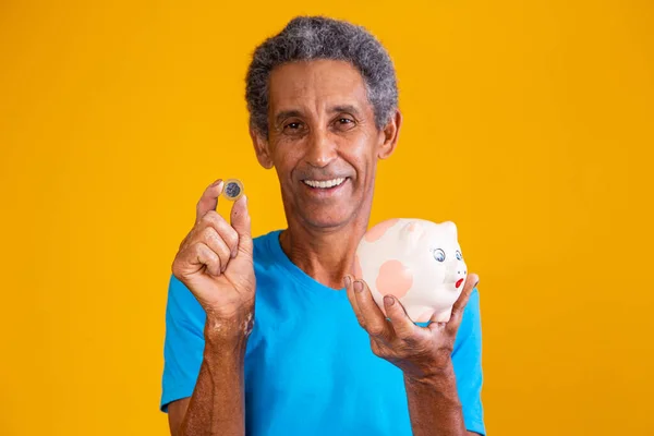 上了年纪的男子拿着一个黄色背景的小猪银行和一枚巴西硬币 财务概念 — 图库照片