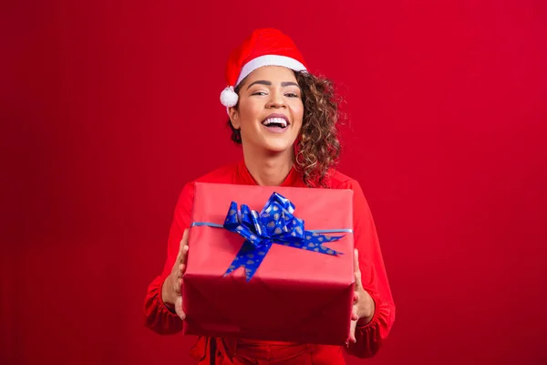 サンタクラスの帽子の若いアフロ女性の肖像画は 赤い背景に贈り物を保持しています クリスマスの夜のコンセプト — ストック写真