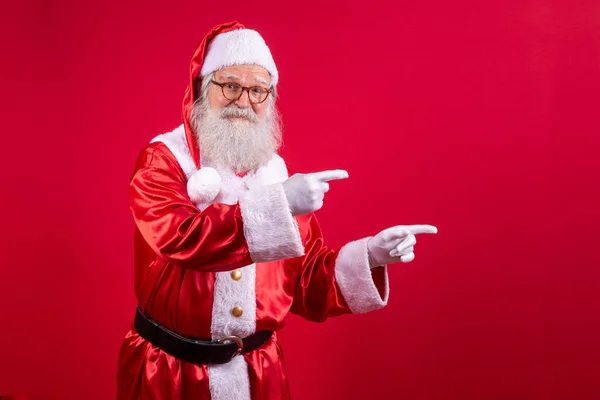 喜形于色的老圣诞老人喜形于色 他身穿盛装 看着镜头 指指点点地将圣诞促销广告放在一边 以红色为背景的圣诞折扣广告 — 图库照片