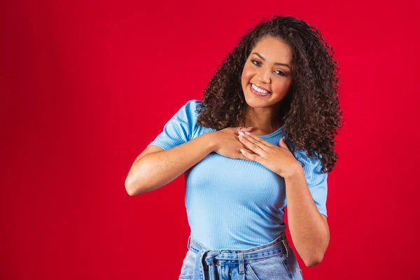 Grateful hopeful happy black woman holding hands on chest feeling pleased thankful, sincere african lady expressing heartfelt love appreciation gratitude honesty isolated on red studio background