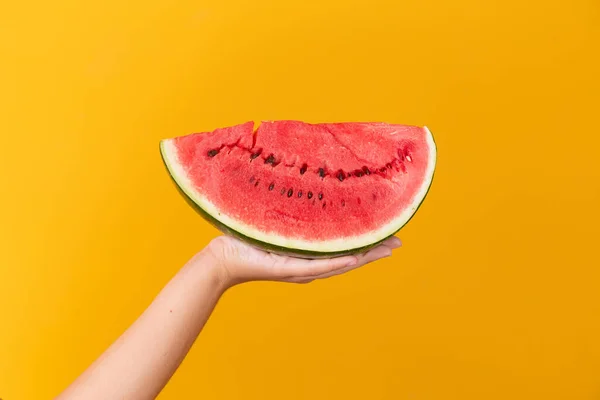 stock image female hand holding a piece of watermelon on yellow background with space for text.