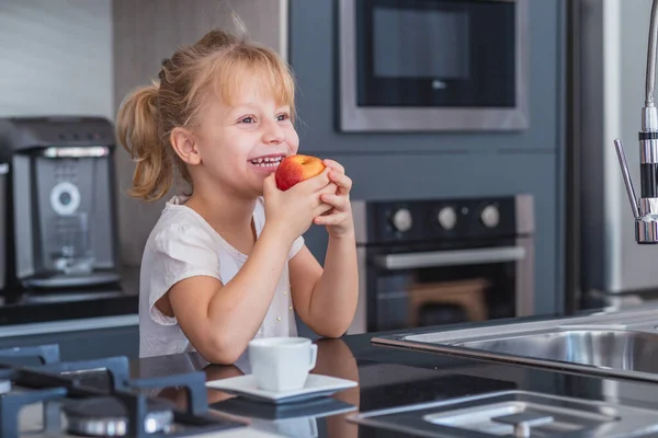 小さなブロンドの女の子が台所でリンゴを食べて — ストック写真