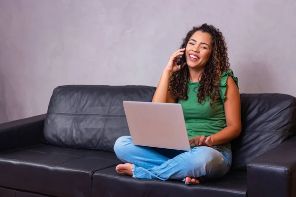 微笑的千禧年非洲裔美国妇女用手机在家庭办公室使用笔记本电脑 快乐的年轻混血女士打电话有手机对话看电脑屏幕坐在坐在屏幕上 — 图库照片