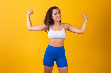 Güçlü ve kendinden emin Afro-Amerikan bir kız görüntüsü, kaslarını esnetiyor ve kendinden emin görünüyor, sarı arka planda duruyor.